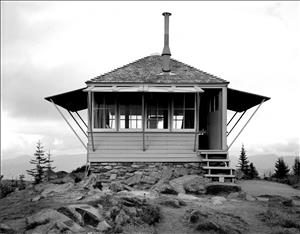 A one room wooden structure with a chimney pipe coming out of the roof. It has windows on all sides and shutters that open upwards. It is perched on a foundation of rocks on a mountain top. Clouds and forest in the distance. 